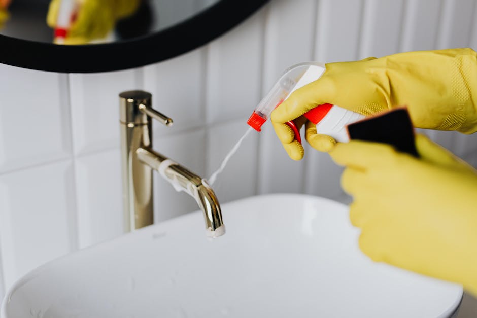 A person wearing yellow rubber gloves is holding a spray bottle filled with cleaning solution and a small black cloth, preparing to sanitise a white ceramic sink with a shiny, stainless steel faucet.